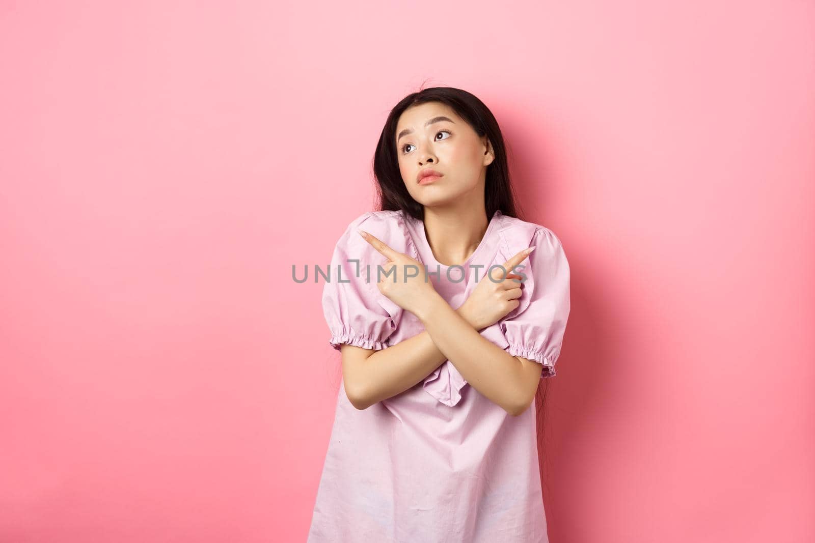 Indecisive asian woman cant decide, pointing fingers sideways and look pensive aside, making choice, standing confused on pink background.