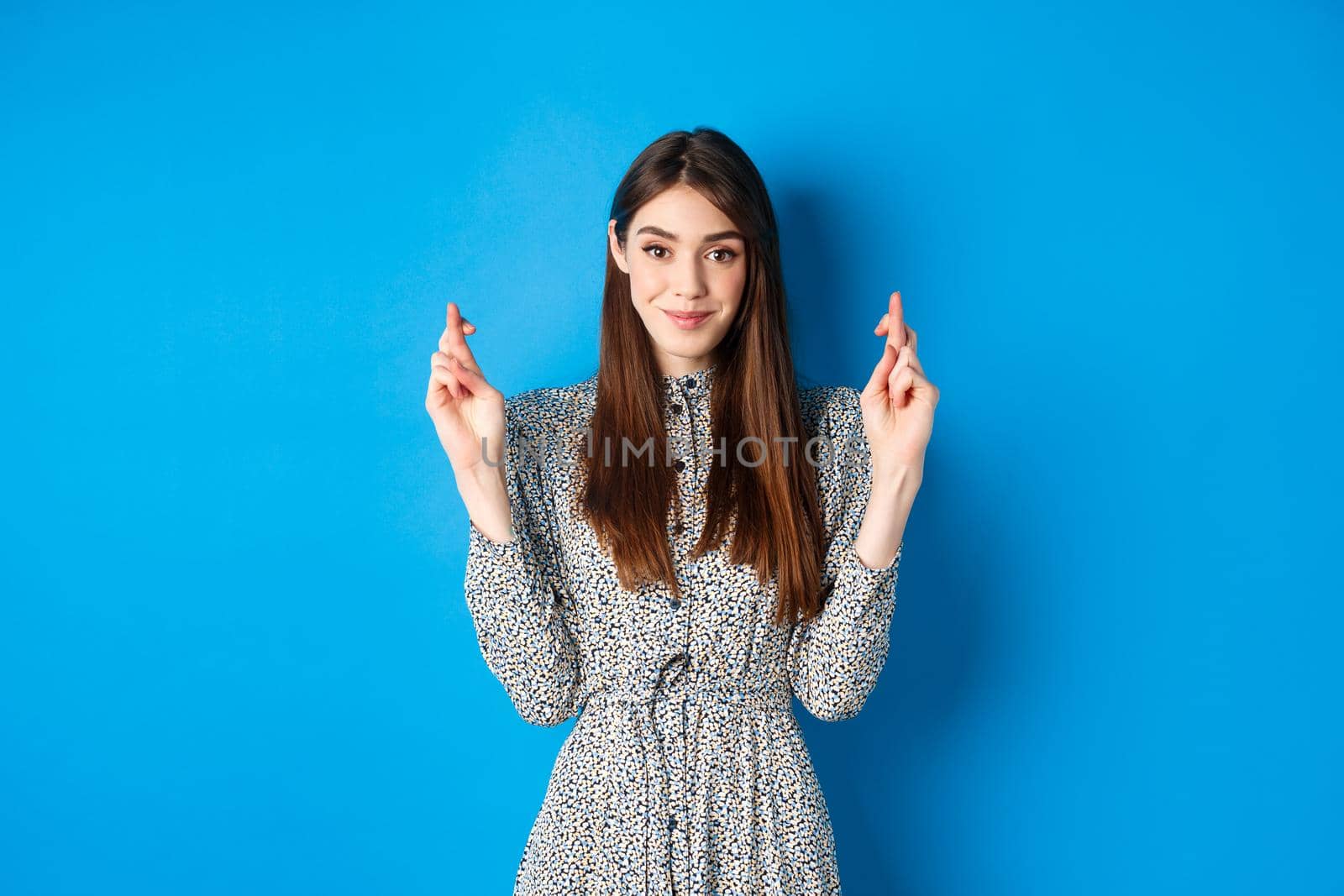 Hopeful smiling woman cross fingers for good luck and looking positive, praying or making wish, standing on blue background.