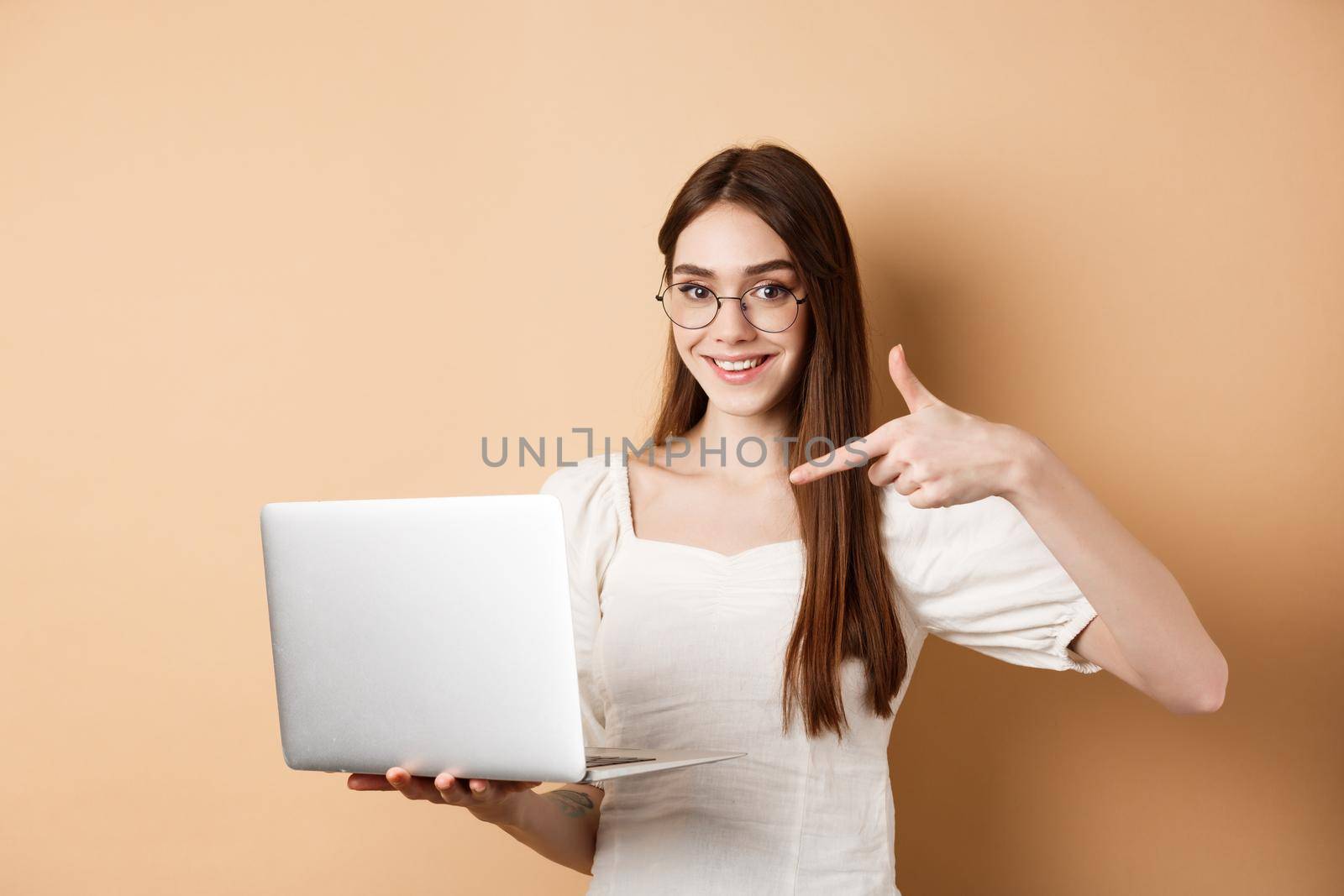 Smiling woman in glasses pointing finger at laptop screen, showing online promo, standing on beige background by Benzoix