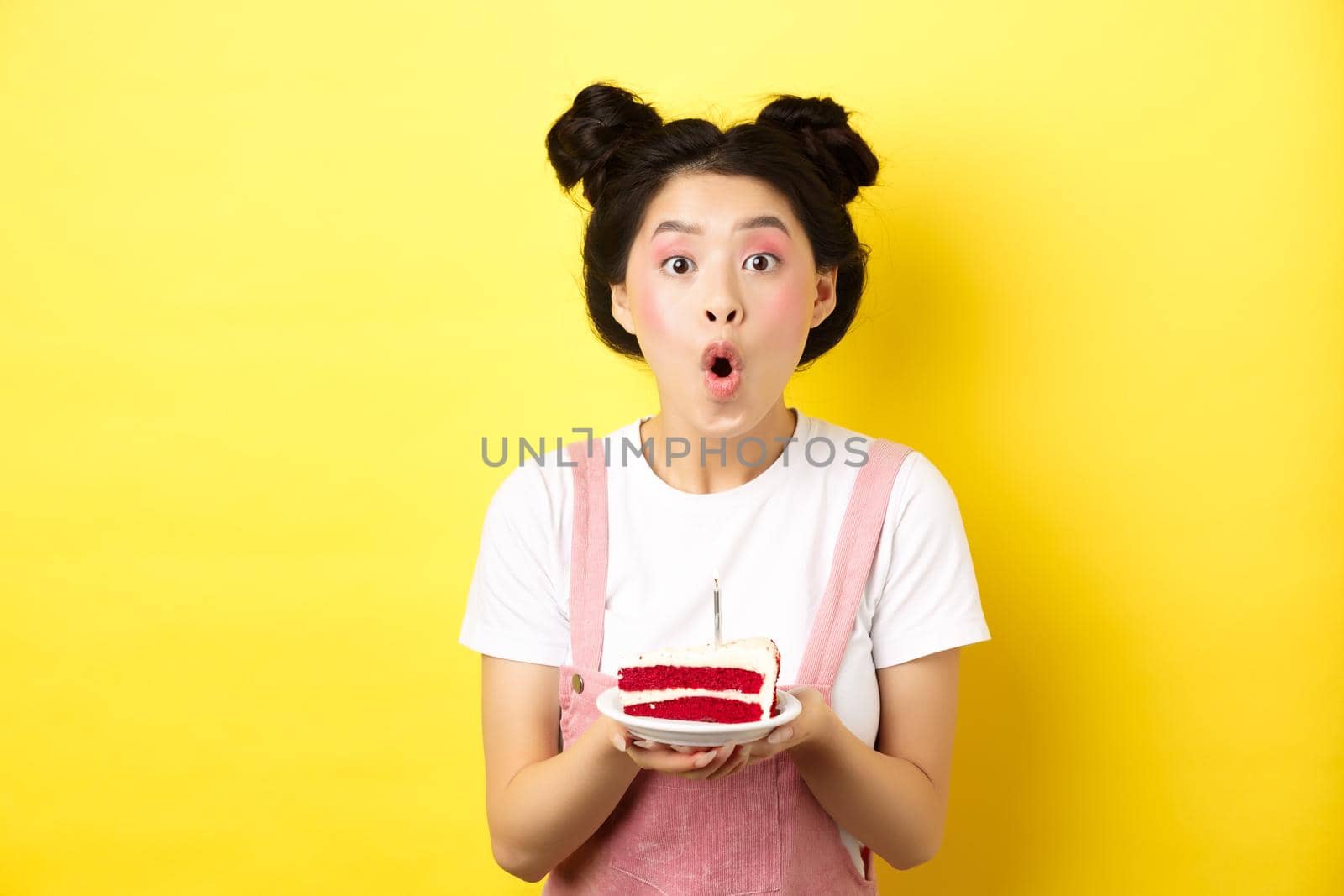 Happy asian birthday girl with bright makeup, blowing candle on cake, making wish, standing on yellow background by Benzoix