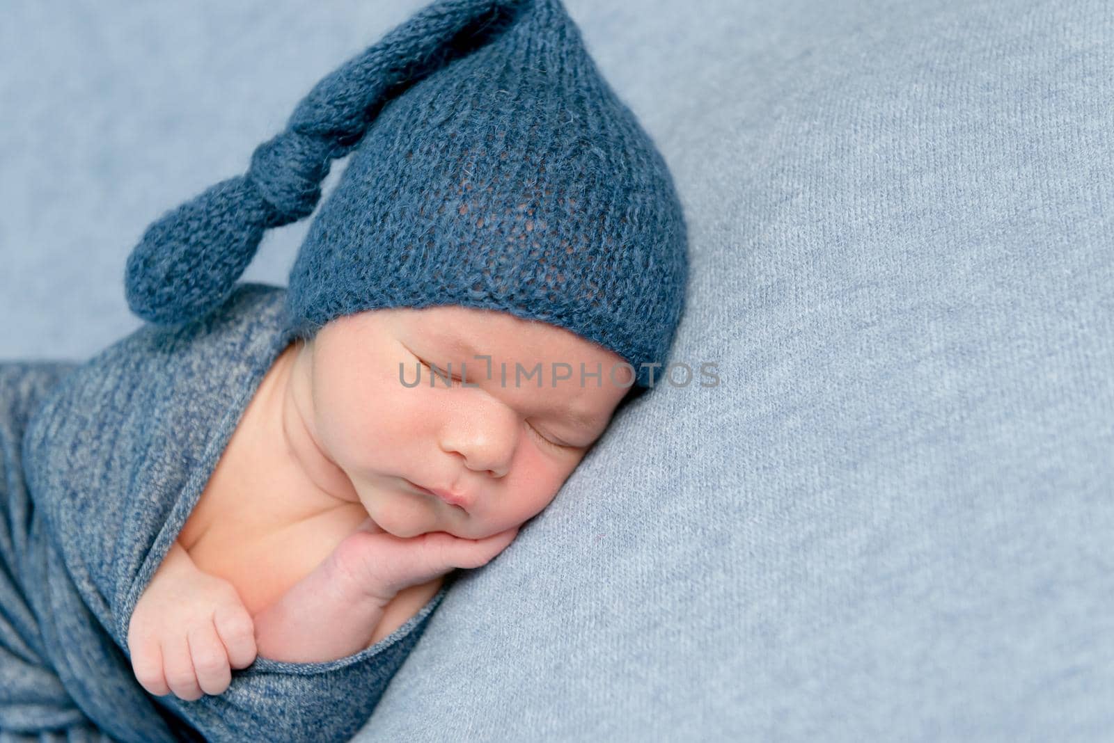 Newborn baby boy swaddled in blue blanket and blue knitted bonnet sleeps peacfully on grey background.