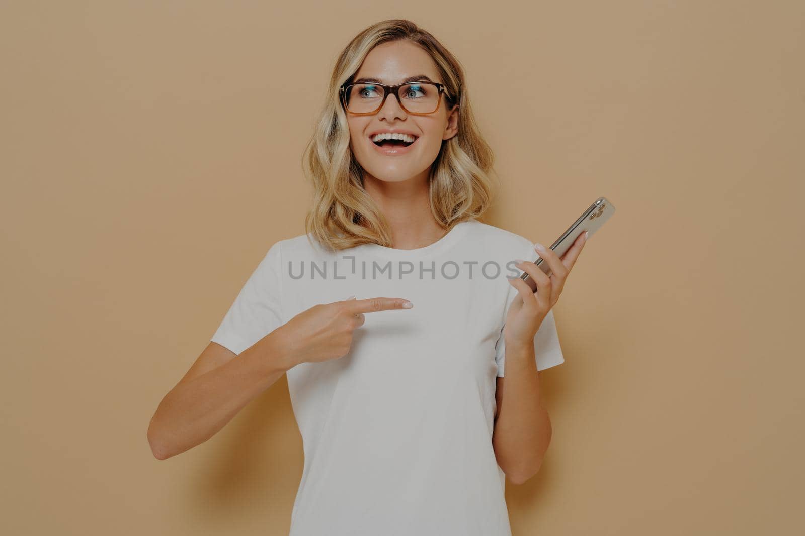 Happy and excited girl in spectacles pointing on mobile phone and looking aside with amazed face expression, reading unbelievable and good news in internet while posing in studio