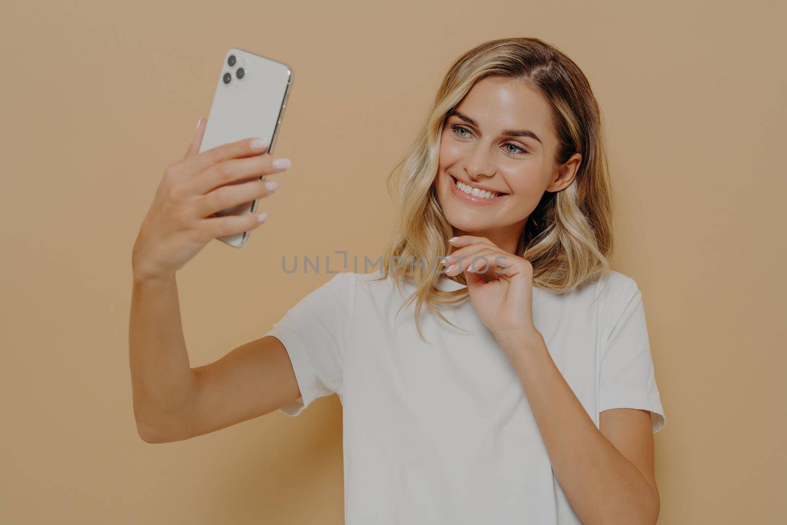 Cute lovely female with blonde hair making selfie on modern mobile phone and smiling, taking picture of herself for boyfriend while posing against nude background in studio. Dressed in white tshirt