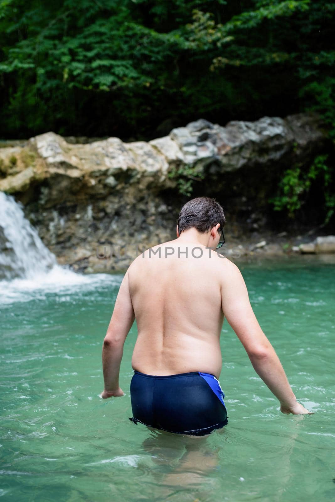 Tropical nature and vacation. Back view of a well-built man swimming in the mountain river with a waterfall