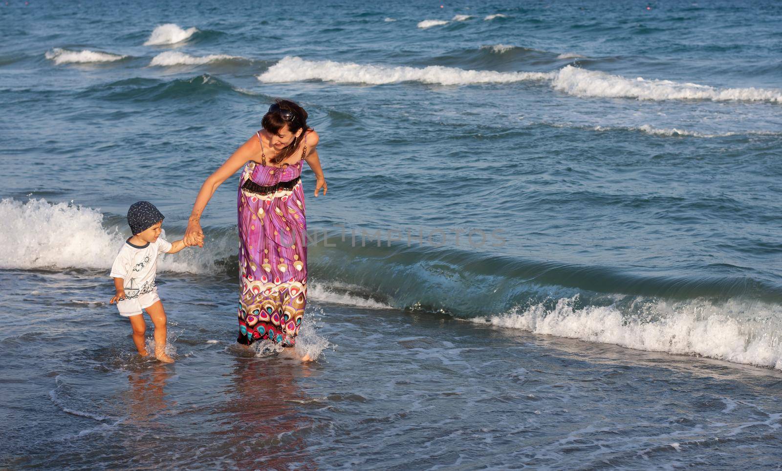 A young woman in a long summer dress walks with her little son among the waves on the seashore