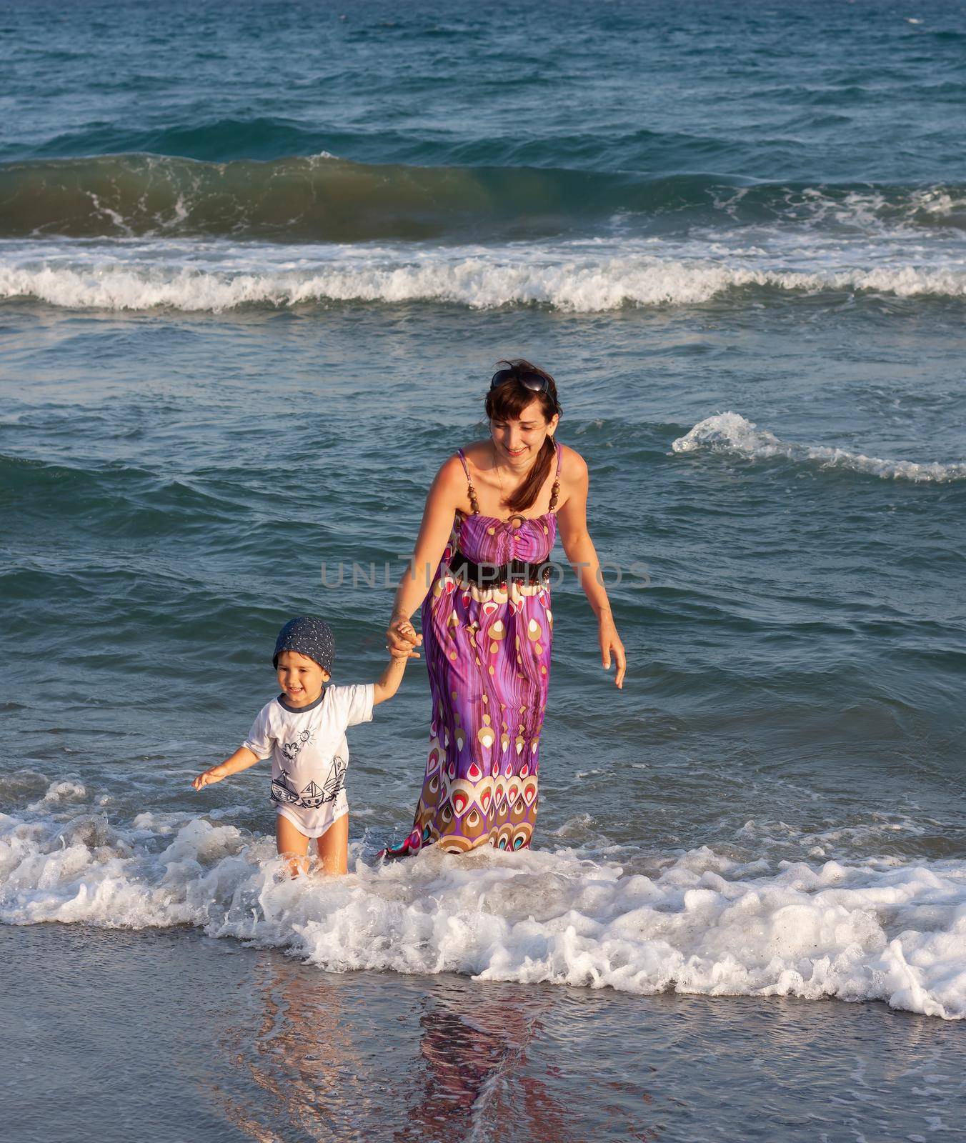 A young woman in a long summer dress walks with her little son among the waves on the seashore