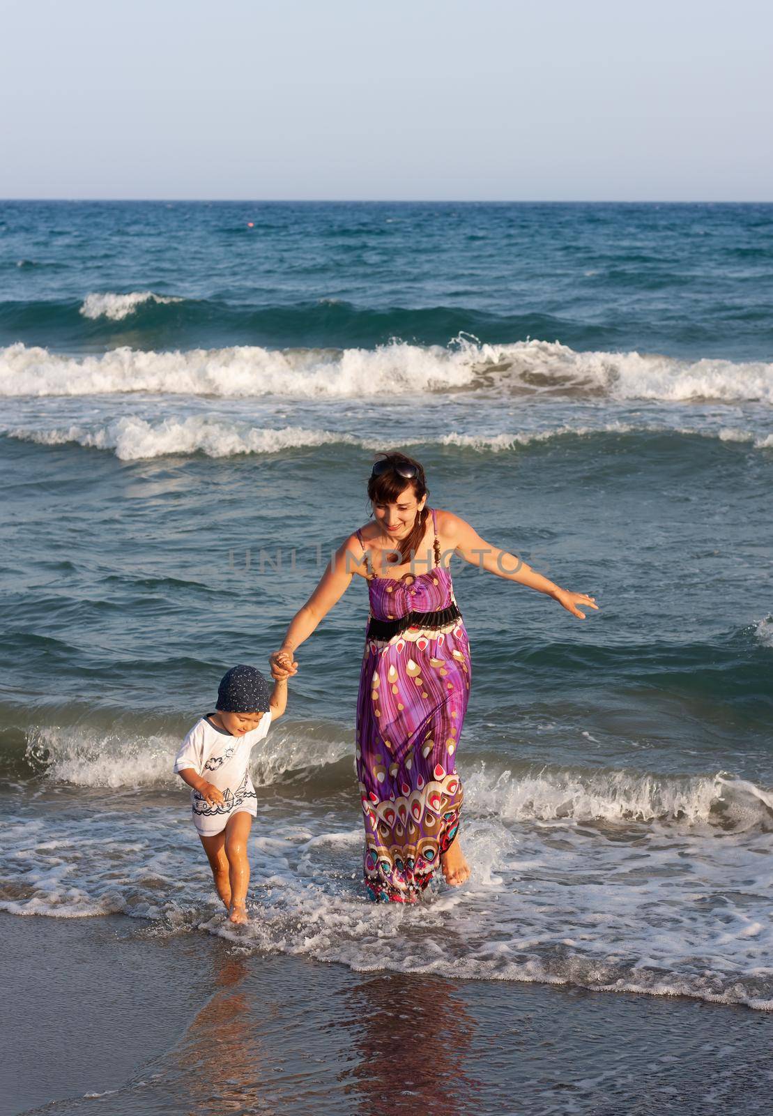 A young woman in a long summer dress walks with her little son among the waves on the seashore