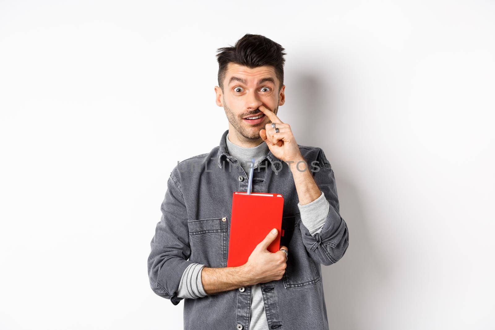 Young guy picking nose and holding book, look stupid at camera, standing against white background.