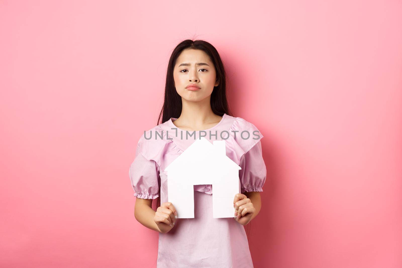 Real estate and insurance concept. Sad frowning asian girl showing paper house cutout and grimacing upset, standing against pink background.
