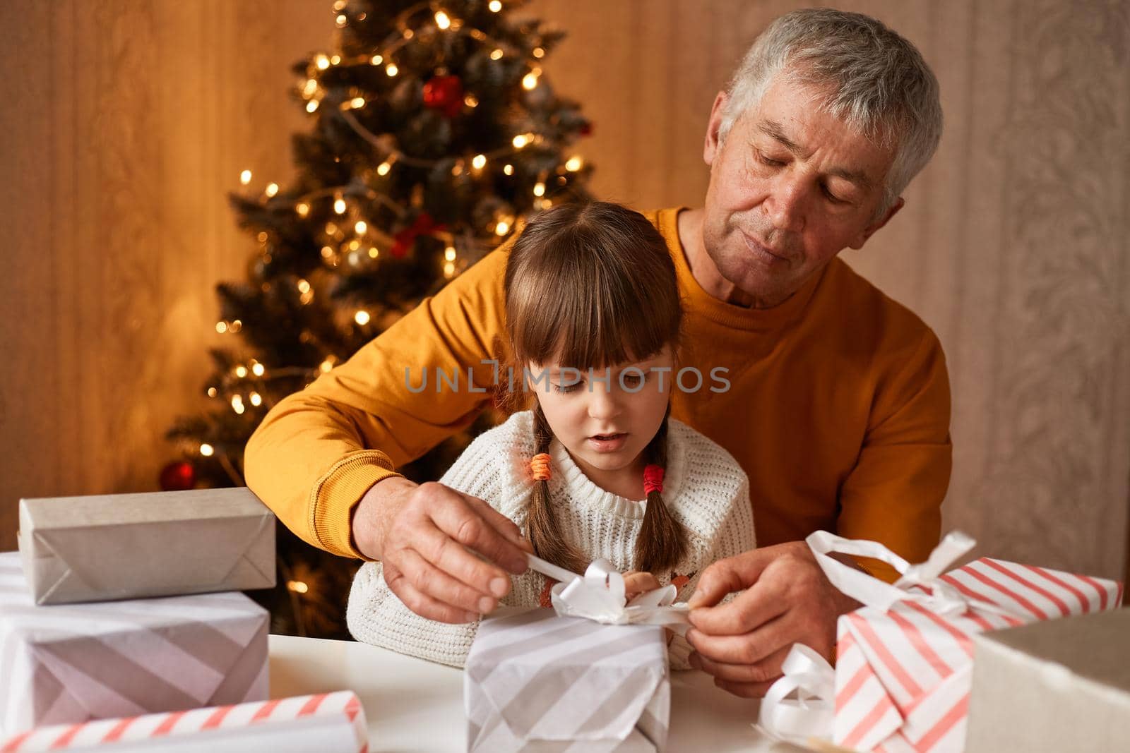 Concentrated little girl with pigtails wearing white sweater posing with her grandfather at table and preparing present boxes for congratulating their family with New year eve.
