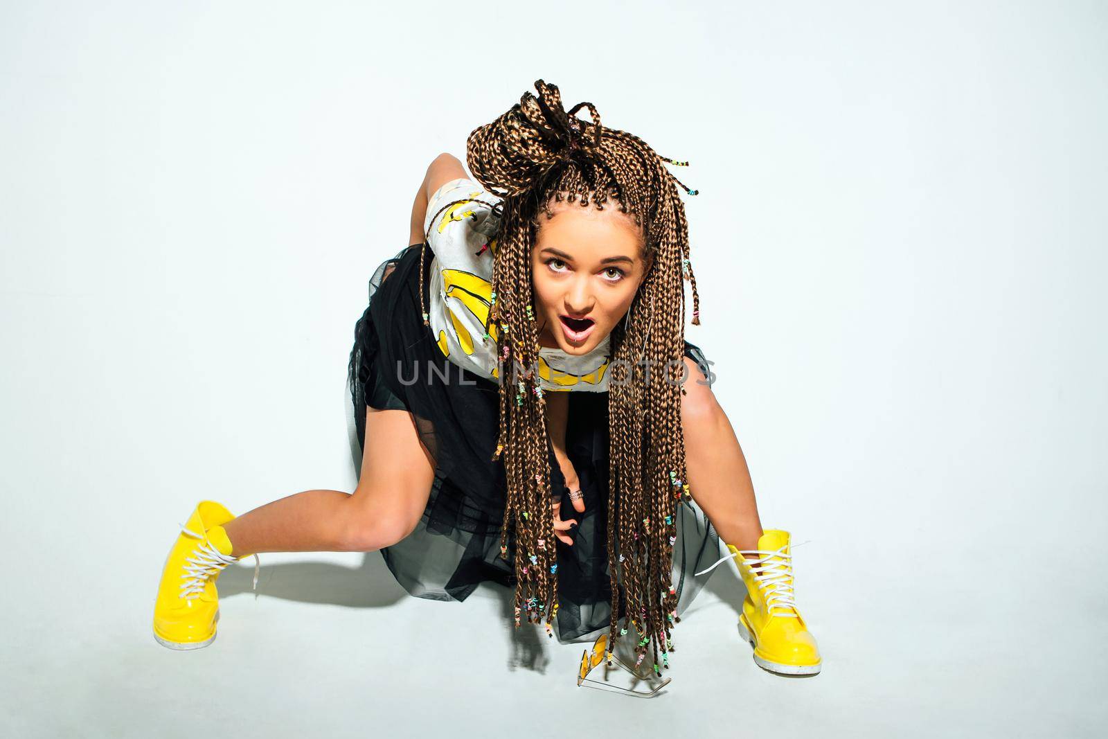 Portrait of a young stylish woman with braided hair dressed in white t-shirt, black skirt, yellow boots and sunglasses posing on the white background.