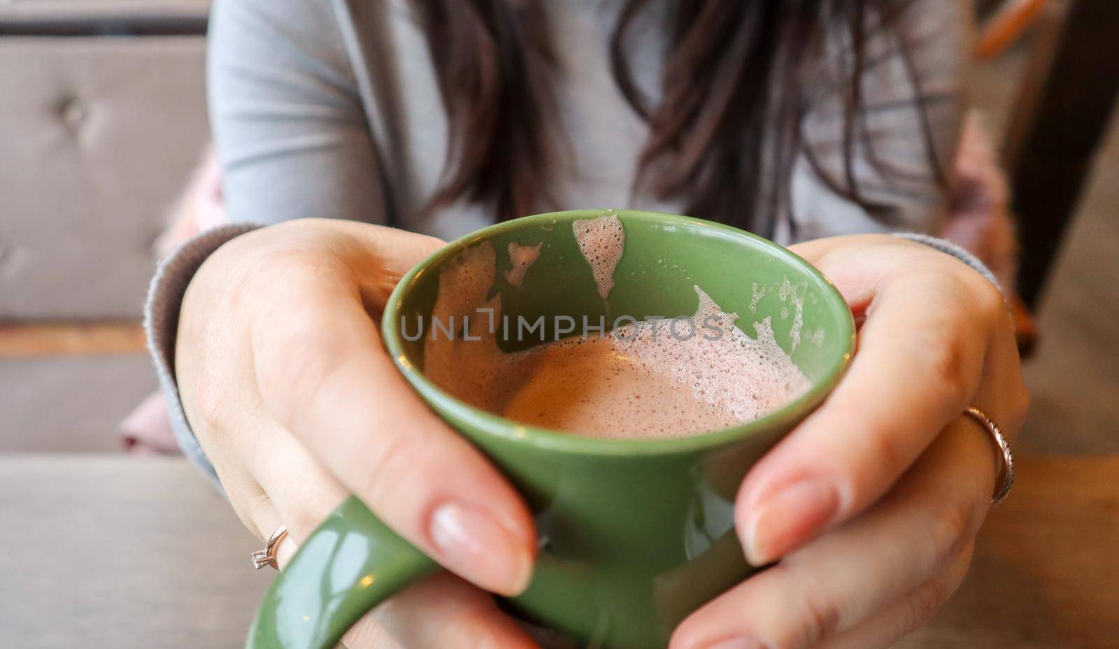 Close up of female hands with a drink mug. A girl in a gray sweater holds a cup of coffee or cocoa in a cafe at the table and looks out the window. Hot drink. A cozy photo with a blurry background.