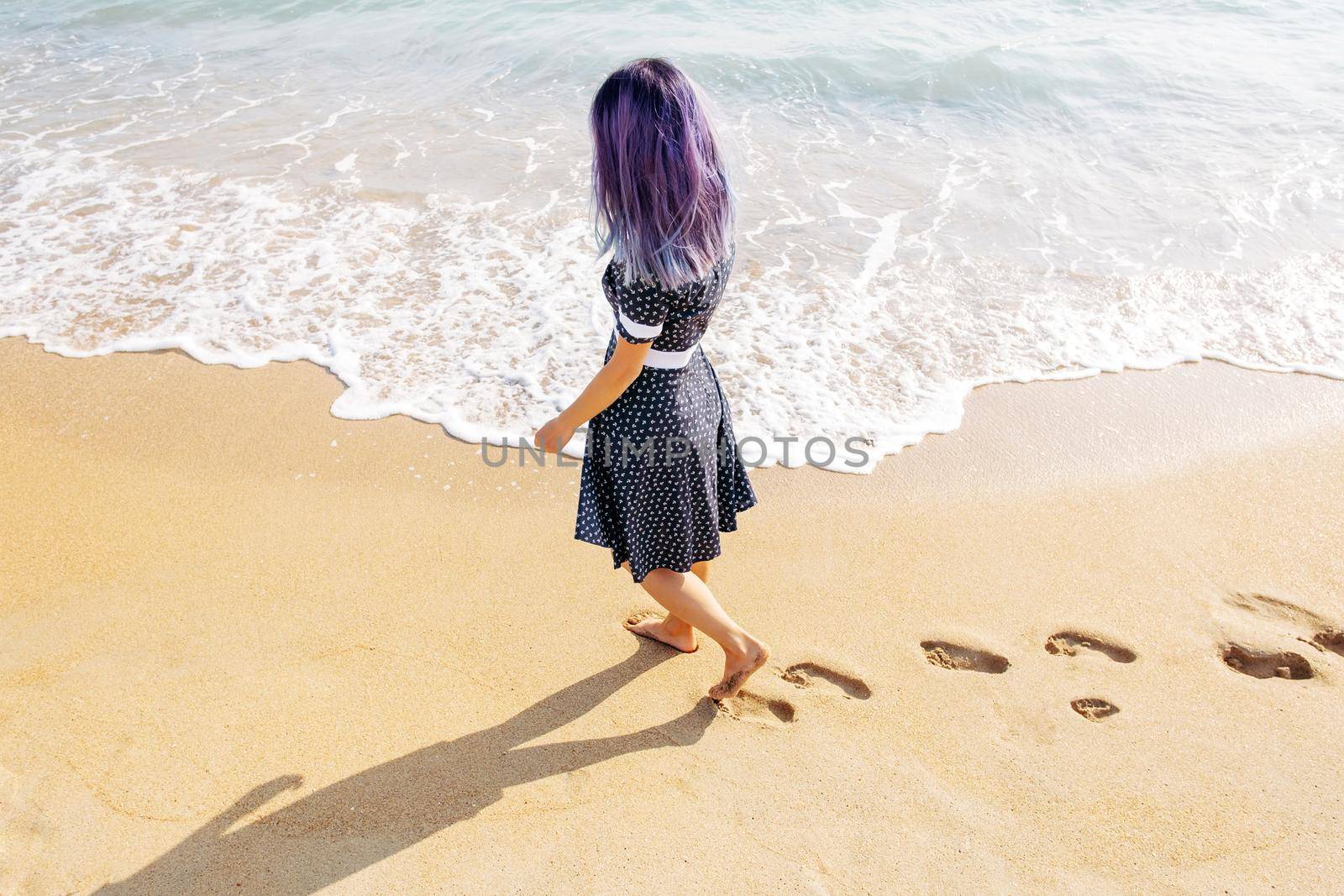 Unrecognizable barefoot young woman walking on beach leaving footprints in the sand near sea. Summer vacations.