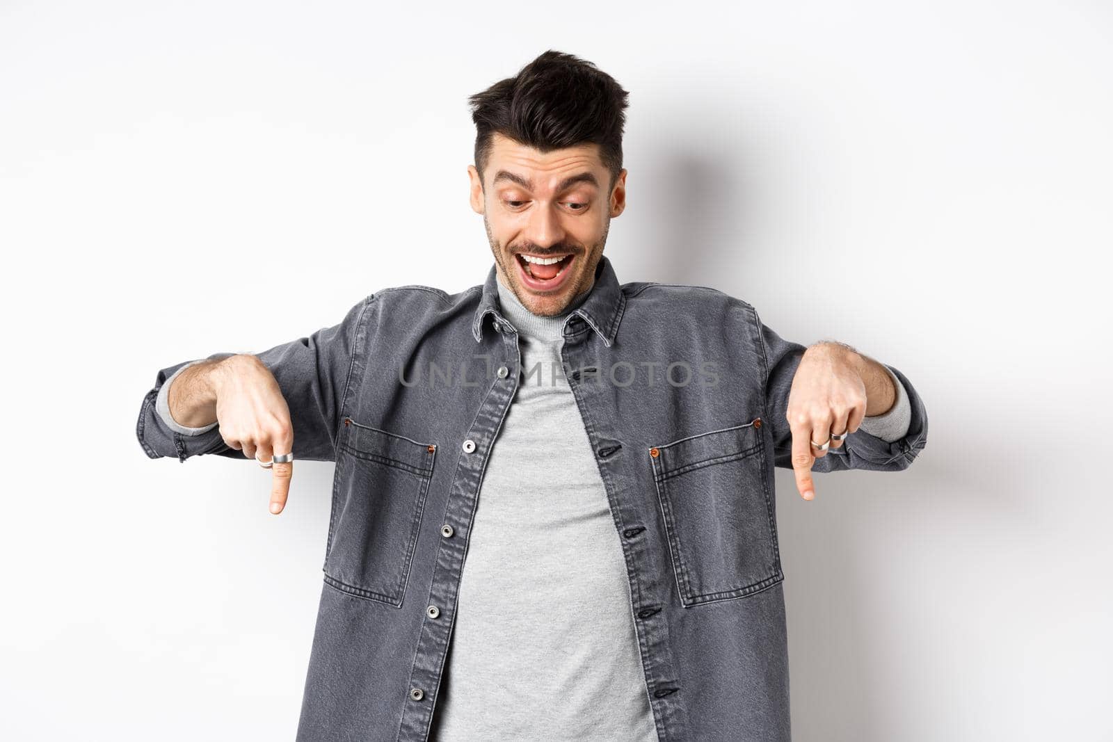 Excited european man looking and pointing fingers down, looking happy at advertisement, standing in denim jacket on white background by Benzoix