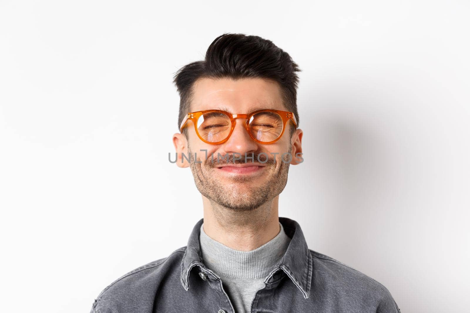 Close-up of cheerful smiling man close eyes, feeling happiness and joy, satisfied with something good, wearing trendy glasses, white background.
