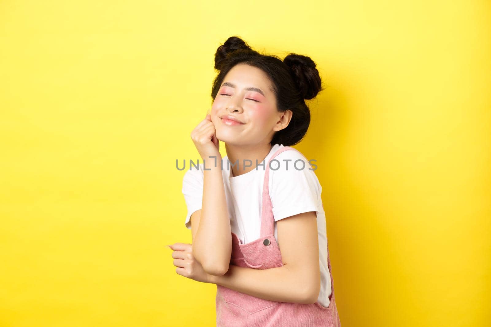 Beautiful happy woman with bright pink makeup, close eyes and touch soft skin, smiling excited, standing in summer clothes on yellow background by Benzoix