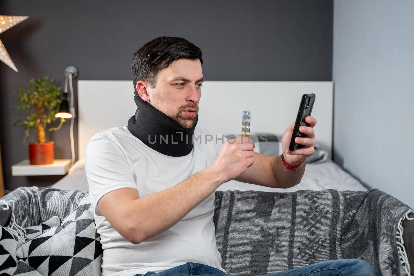 Patient with collar and neck injury receives an online video consultation with doctor and holds pill in hand. Man in foam cervical collar calls traumatologist on video and shows medications on phone by Tomashevska