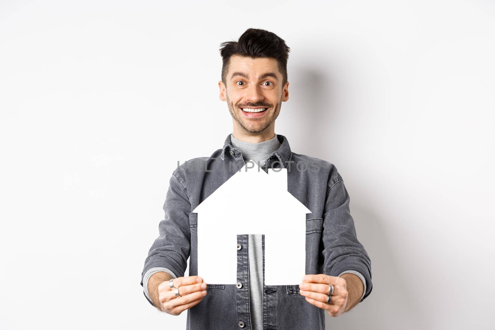 Real estate and insurance concept. Handsome young guy showing paper house cutout and smiling, searching for apartment, buying property, standing against white background by Benzoix