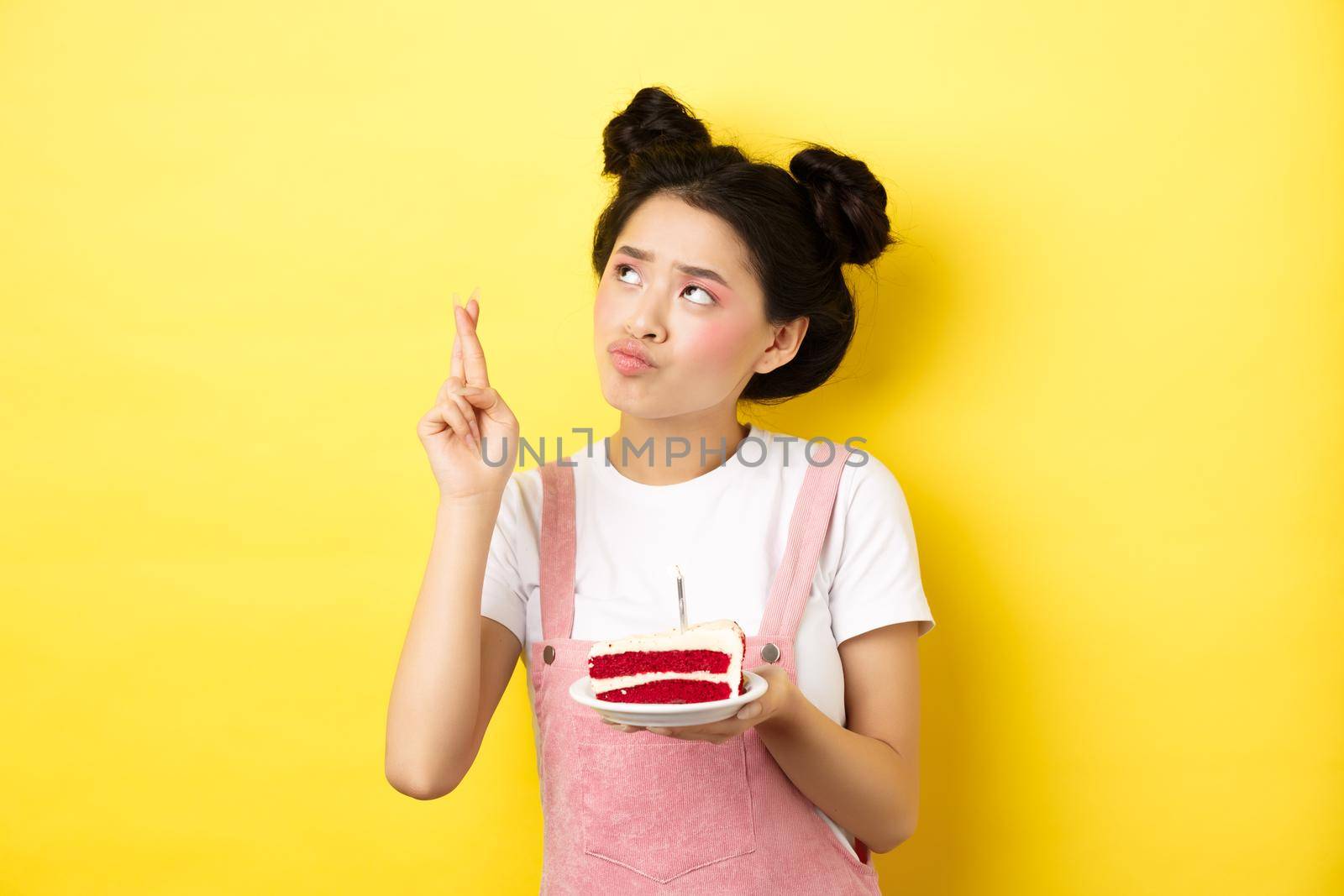 Holidays and celebration. Cute asian birthday girl making wish at party wish cake and candle, cross fingers and looking up, standing on yellow background.