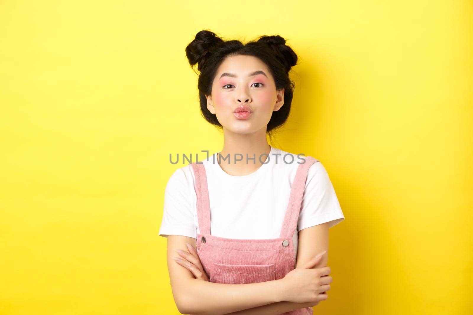 Cute teen asian girl pucker lips for kiss, cross arms on chest and look romantic at camera, yellow background by Benzoix