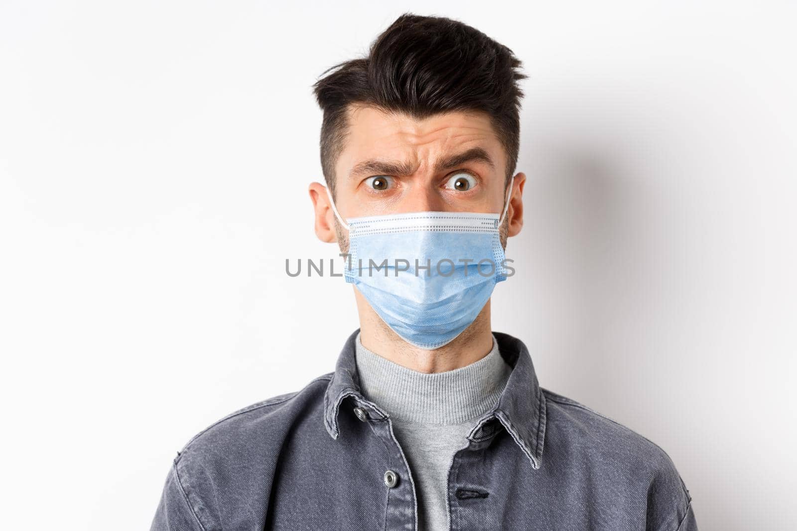 Covid-19, health and lifestyle concept. Confused man in face mask raise eyebrow puzled, cant understand something, standing against white background.