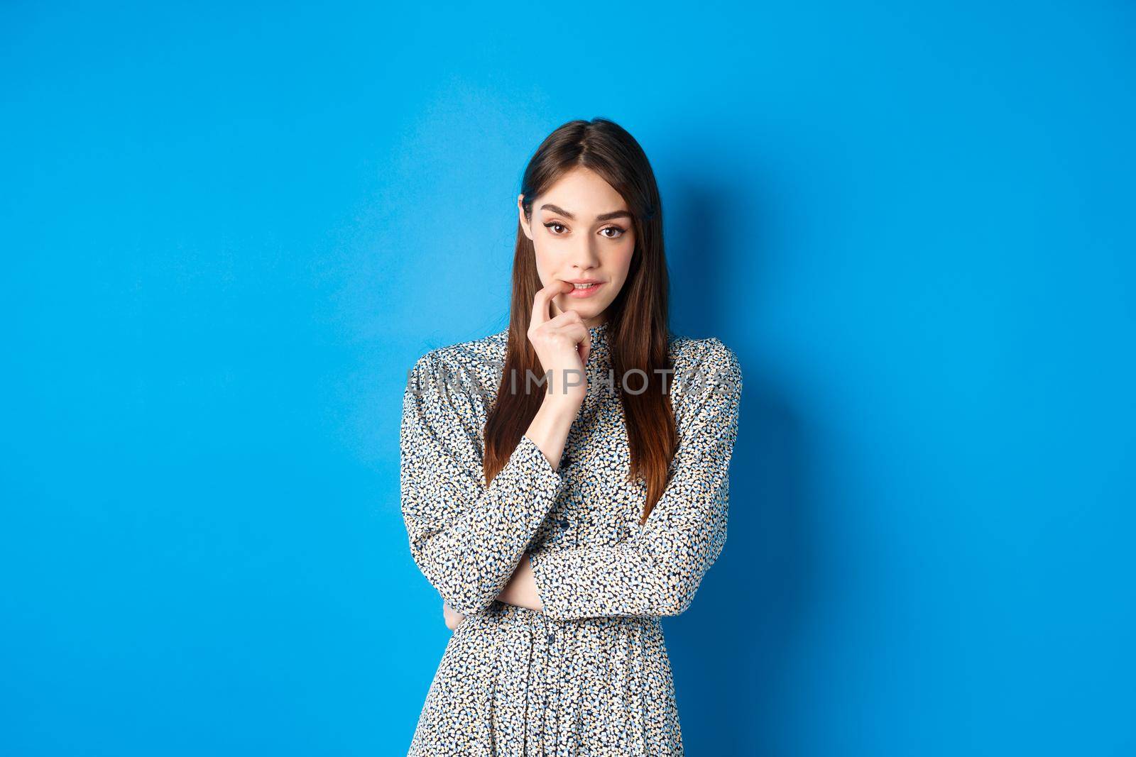 Sensual woman in dress biting finger and looking pensive at camera, standing dreamy against blue background by Benzoix