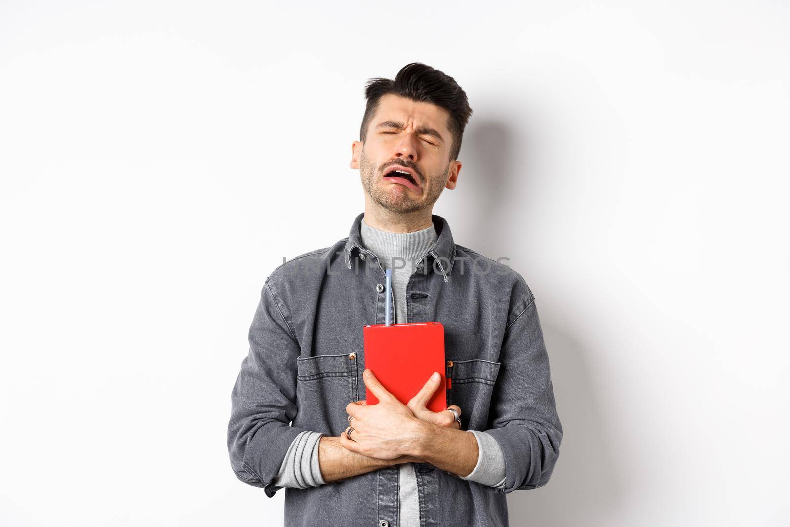 Sad cryig man holding red diary and sobbing, miserable guy carry journal with him, standing against white background.