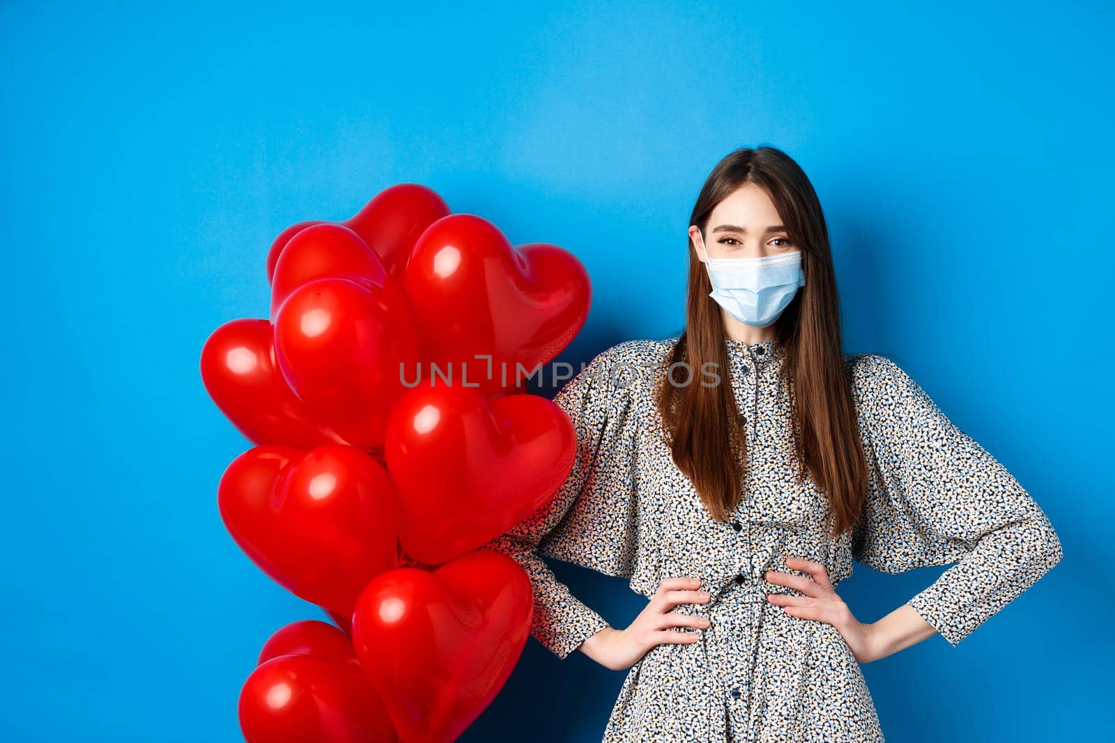 Pandemic and Valentines day. Cheerful smiling girl in medical mask, standing near romantic heart balloons and looking at camera, wearing dress, blue background.