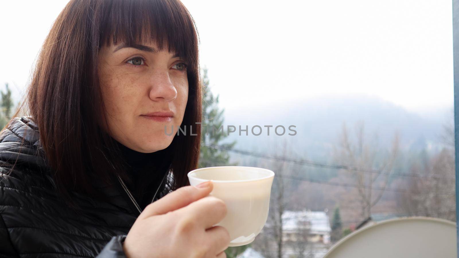 Beautiful happy woman with a white cup of coffee or tea enjoying the beautiful view of the mountain landscape in the morning fog from the balcony of her hotel room. Autumn and winter travel concept by Roshchyn