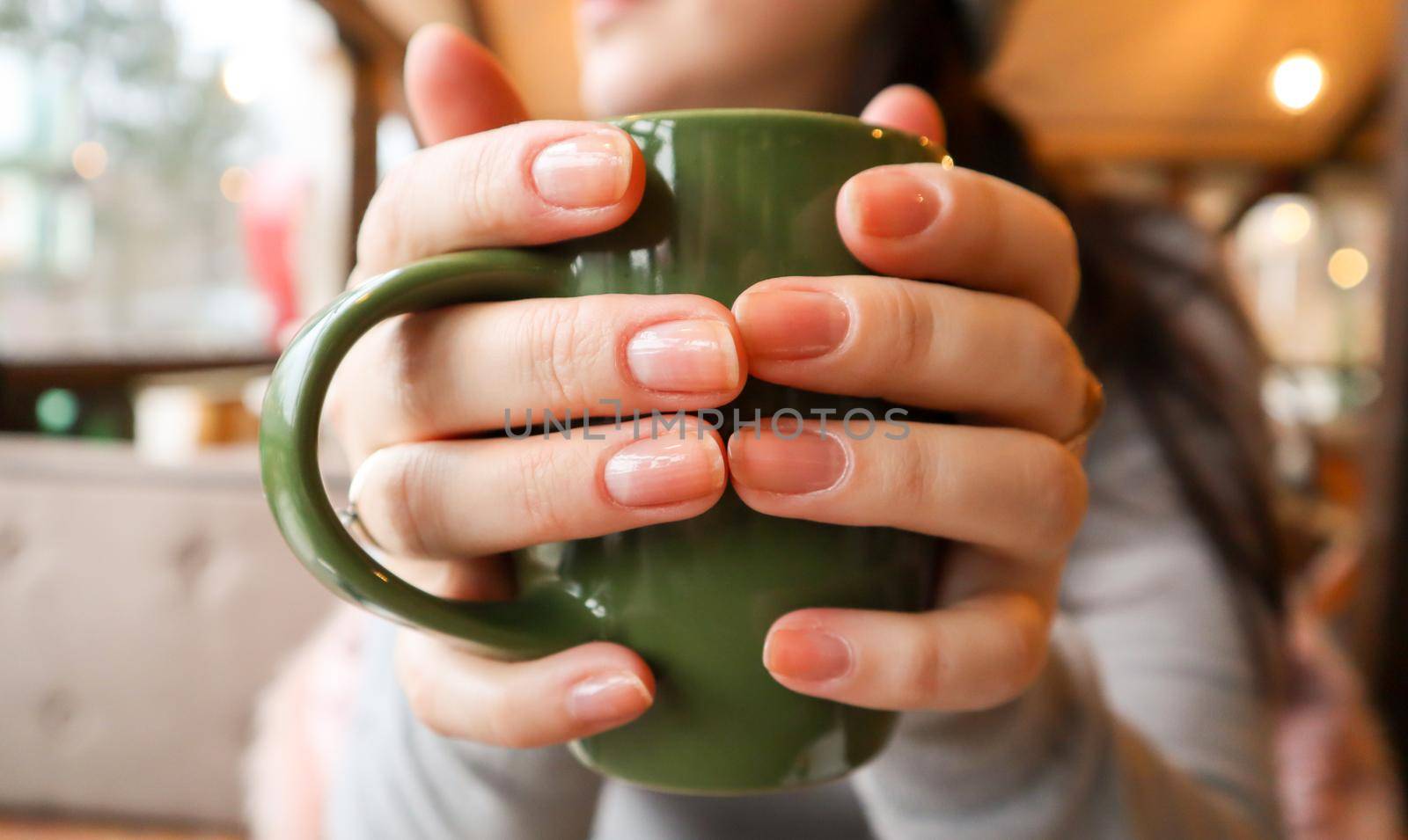 Close up of female hands with a drink mug. A girl in a gray sweater holds a cup of coffee or cocoa in a cafe at the table and looks out the window. Hot drink. A cozy photo with a blurry background.