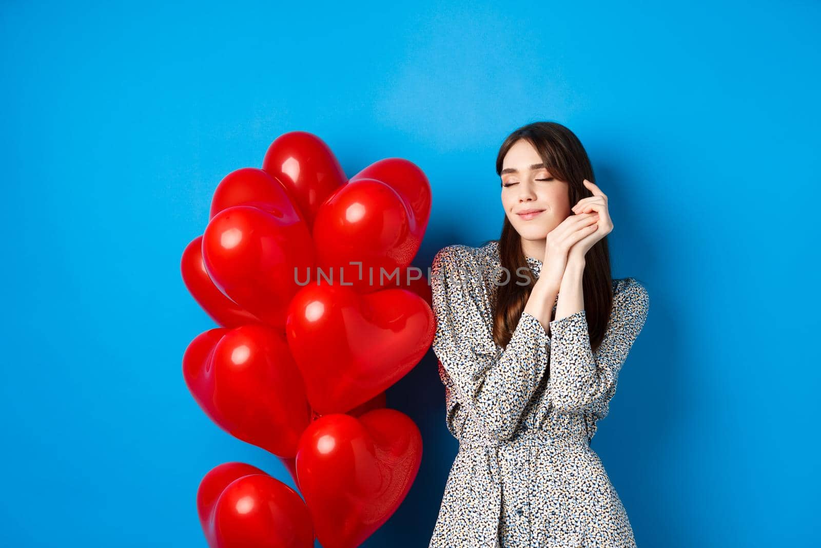 Valentines day. Dreamy romantic woman close eyes and imaging lovely date, standing near heart balloons and smiling, blue background.