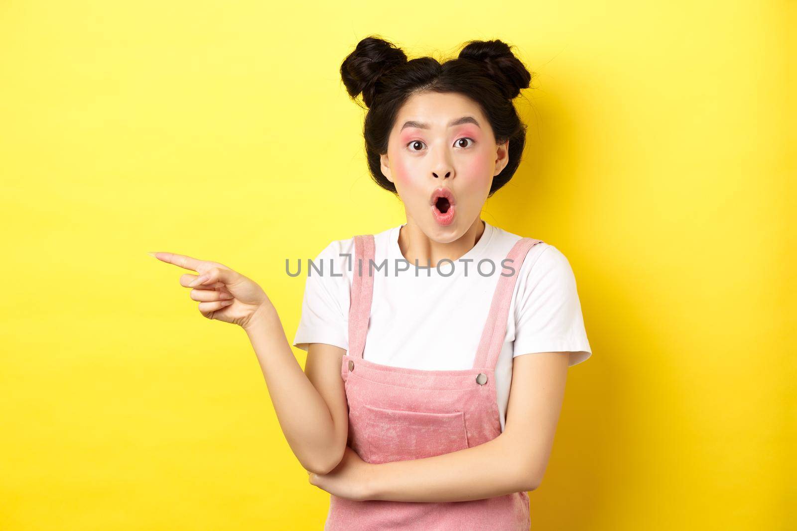 Surprised asian teen girl looking impressed, say wow and pointing finger left, standing with glamour makeup and summer clothes on yellow background by Benzoix