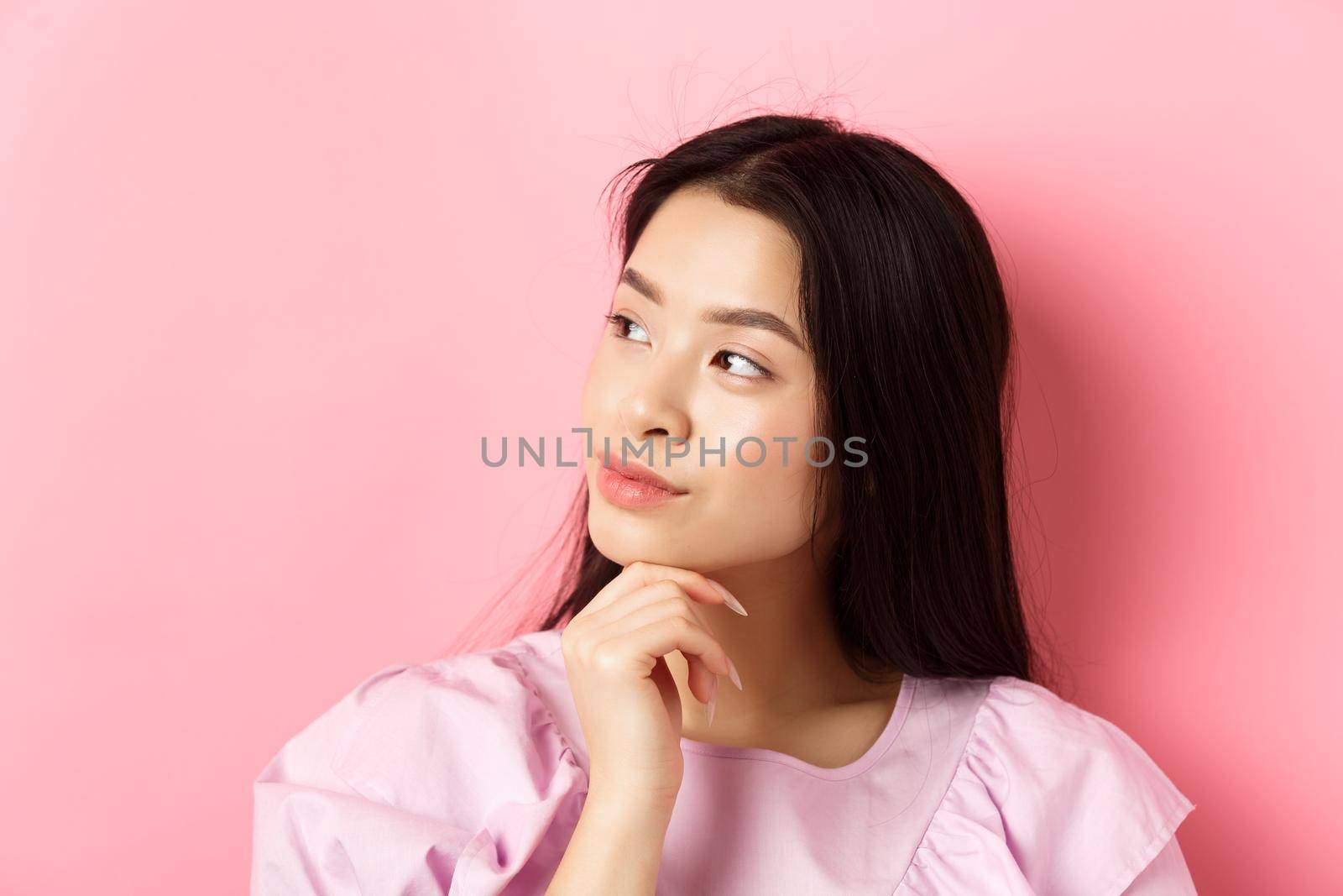 Close-up portrait of romantic asian girl looking aside at logo, thinking or daydreaming, standing against pink background by Benzoix