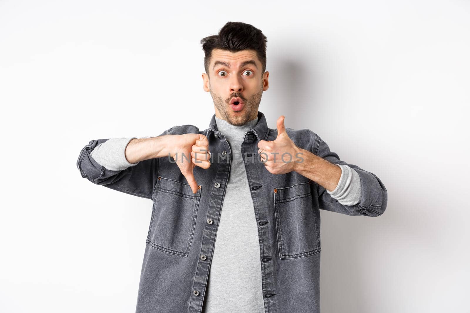 Excited guy show thumbs up and down judging and making decision, rate something interesting, standing on white background by Benzoix