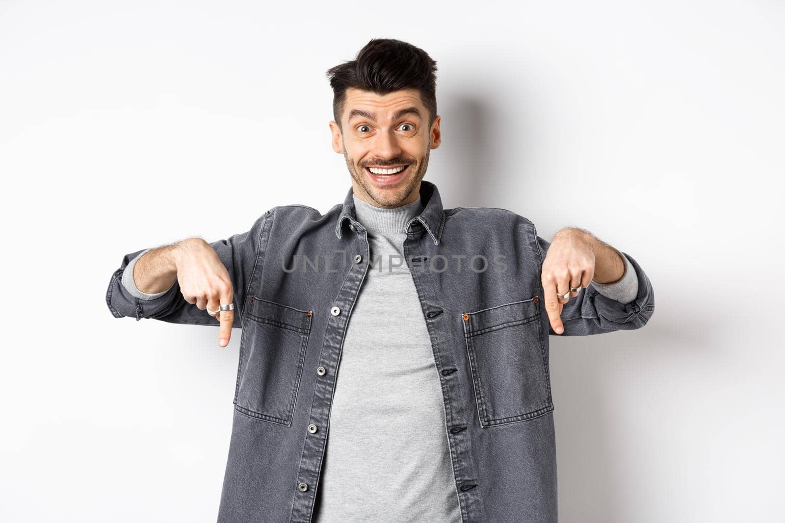 Excited smiling male model pointing fingers down, showing logo with happy face, demonstrate cool promo, standing on white background.