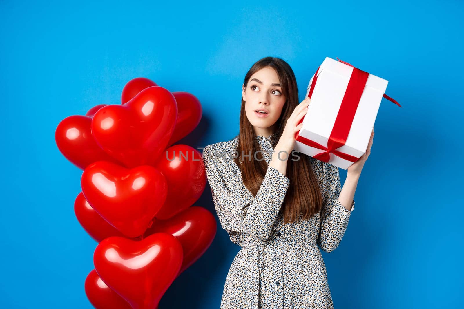 Valentines day. Beautiful woman shaking gift box to guess what inside, look dreamy, celebrating lovers holiday, standing near red hearts, blue background.