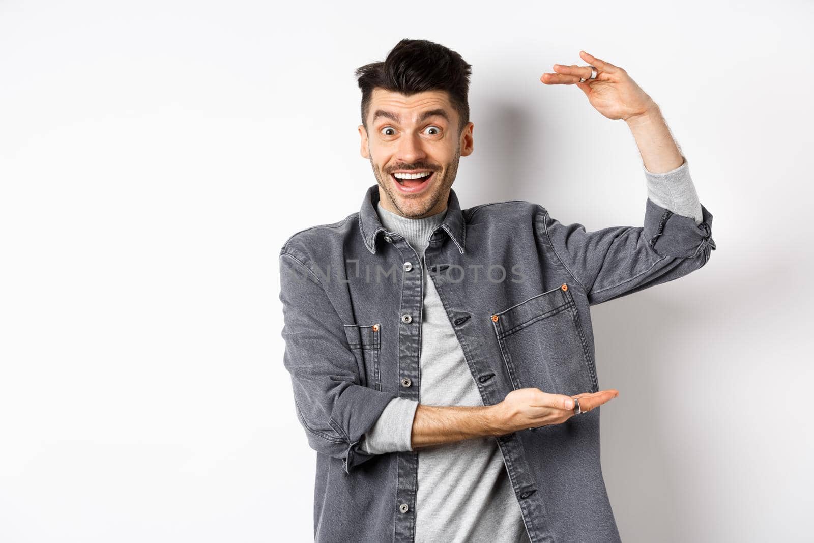 Cheerful guy smiling and showing big size, showing large thing and looking excited, standing against white background.