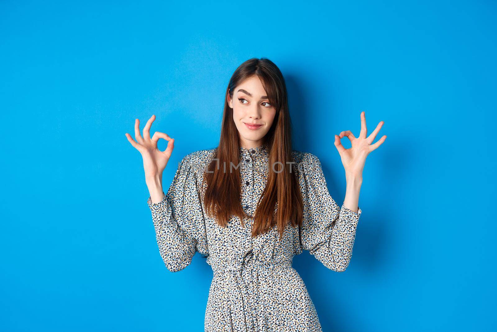 Not bad. Pleased smiling woman with long hair, wearing dress, showing okay signs and looking satisfied, praising good job, looking left at logo, blue background by Benzoix