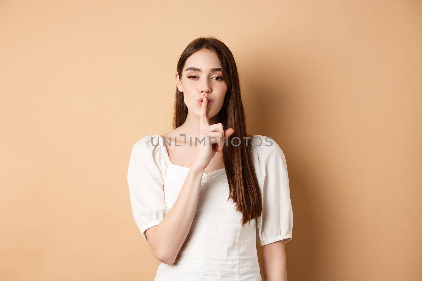 Sensual cute girl hiding secret, showing hush gesture, make shhh sound with finger on lips, stay quiet sign, standing on beige background by Benzoix