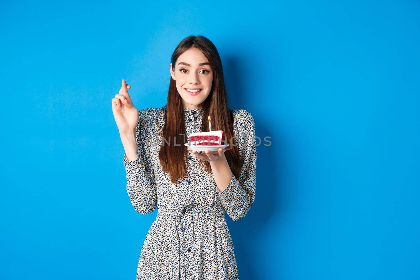 Hopeful birthday girl making wish on cake, cross fingers for good luck, standing in stylish dress against blue background.