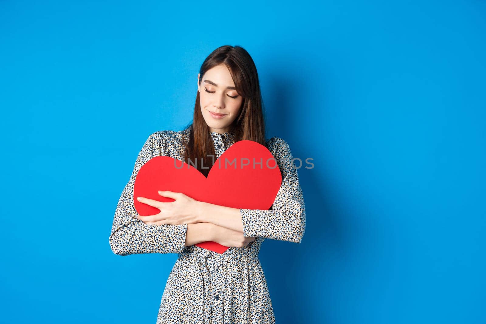 Valentines day. Romantinc girl in dress hugging big red heart cutout, close eyes and smile with dreamy face, imaging sensual date, standing on blue background by Benzoix