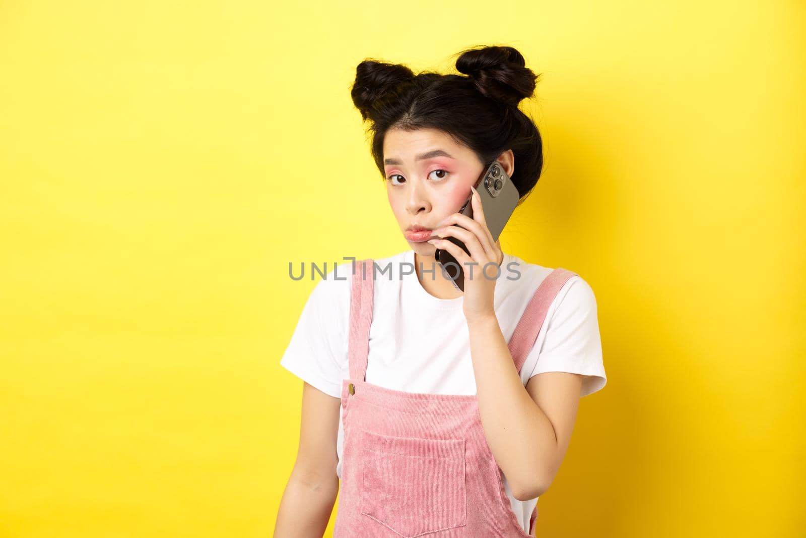 Cute teen girl talking on smartphone, making silly pouting face and look timid at camera, standing with glamour makeup on yellow background by Benzoix