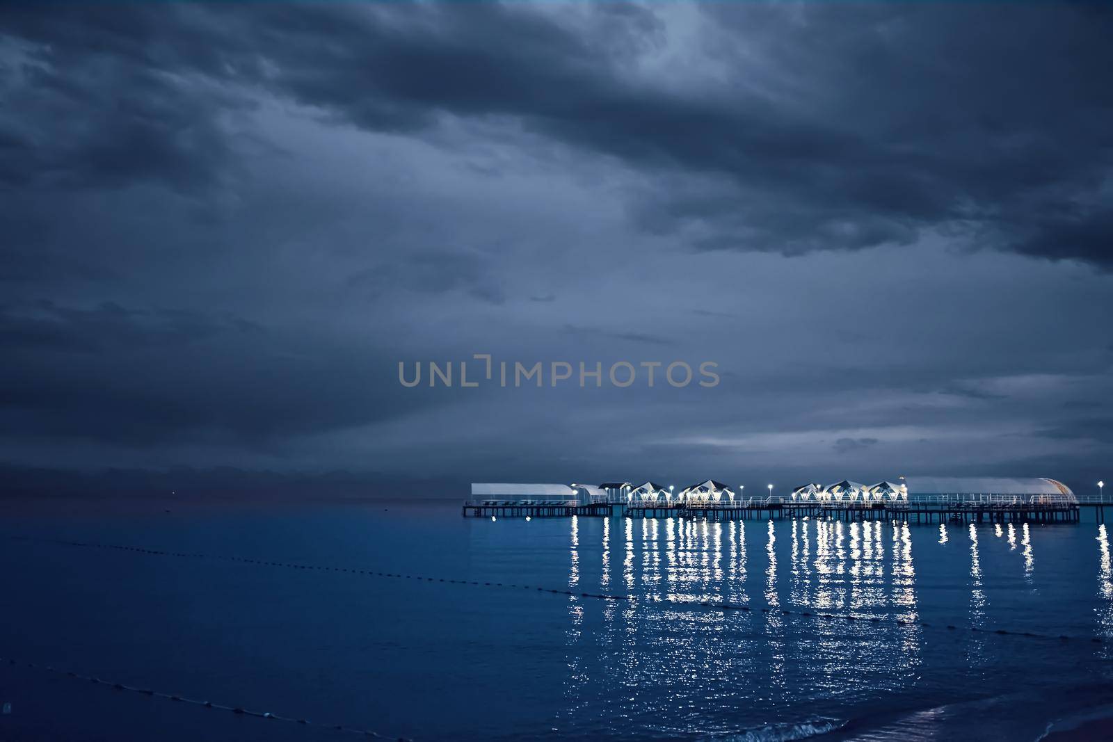 Twilight landscape of pier stretching out into sea. Cloudy sky. Illumination and umbrellas on ocean bridge. Nature summer landscape. Tourism background. Travel and vacation. Glare from lamps on water.