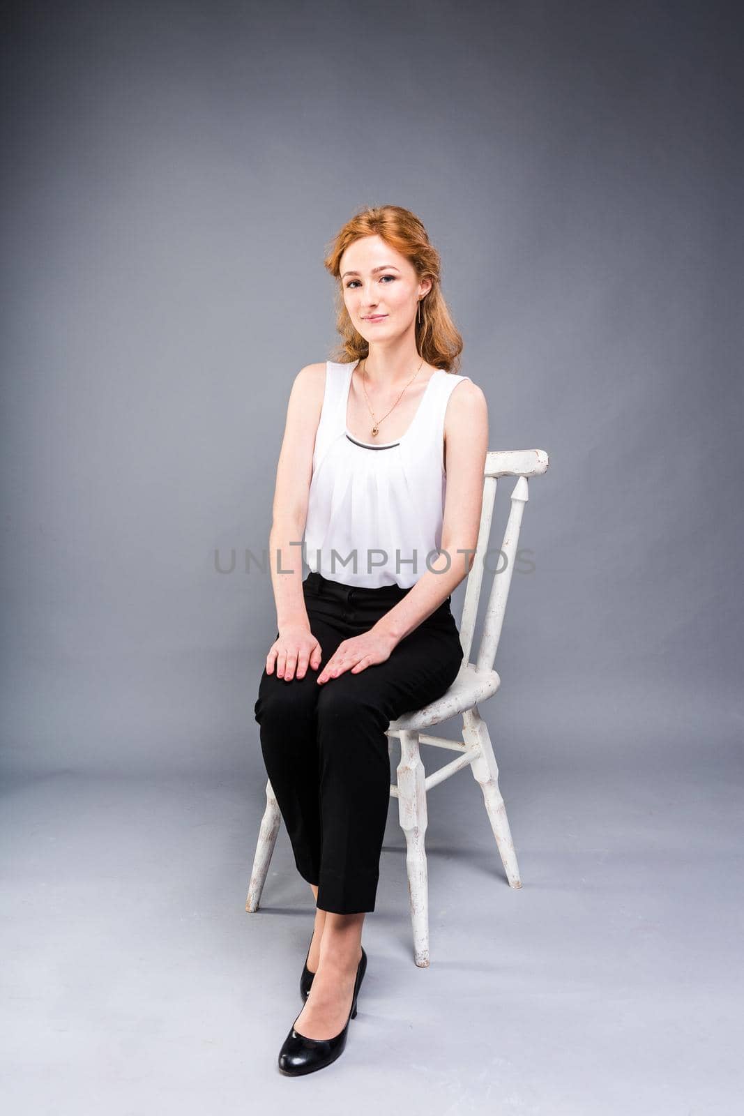 beautiful young caucasian woman with long red hair in high heels sitting on a chair, black trousers and white shirt in full growth on a gray background. Business concept by Tomashevska