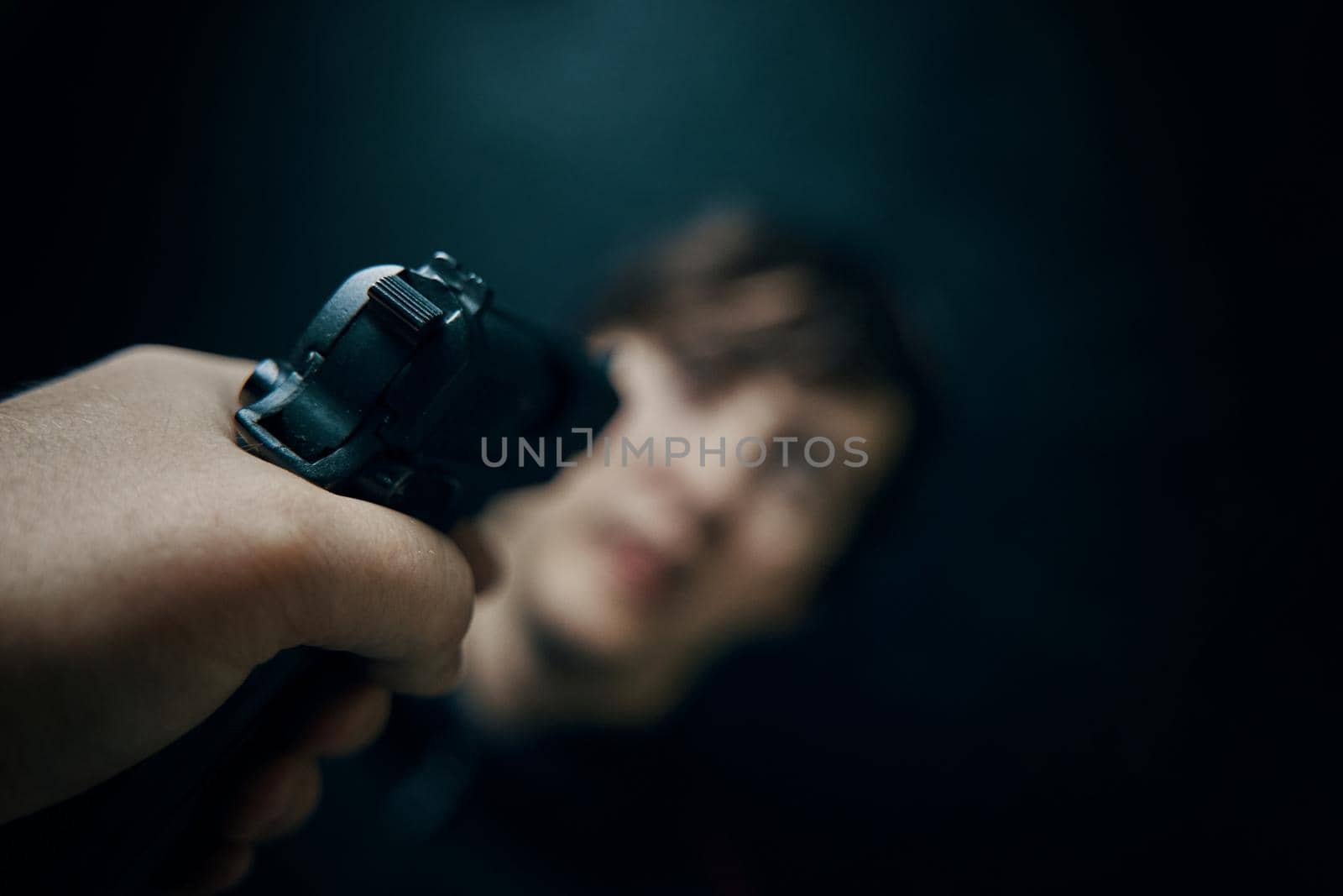 First person view of pistol aimed at young man on dark green background. Firearm in man's hand. POV of aiming at human target. Criminal with gun. Murderer or armed thief.