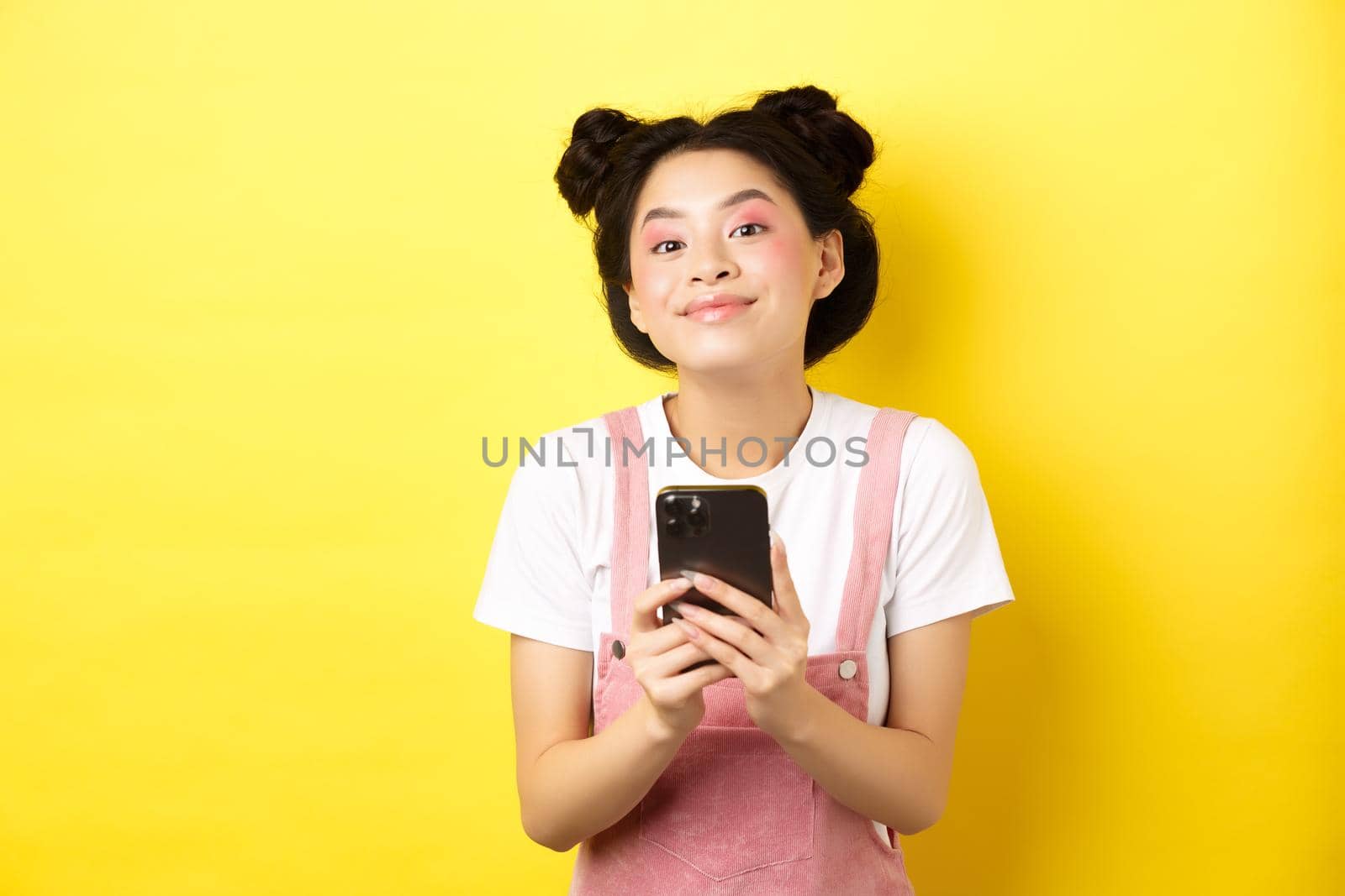 Cute stylish asian girl using mobile phone, wearing glamour pink makeup and summer clothes, standing on yellow background.