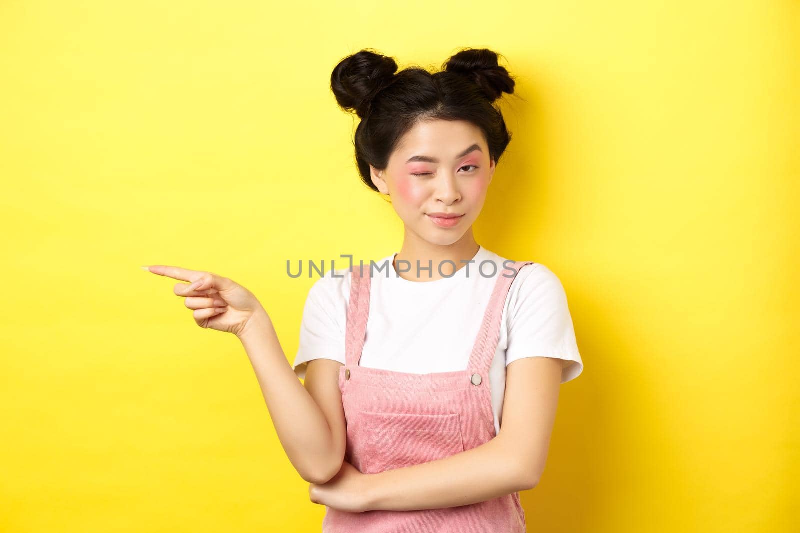 Glamour asian girl with bright makeup, pointing finger left and winking at camera, hint on good promo deal, standing on yellow background.