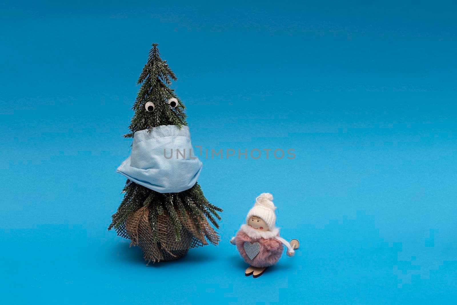 Christmas tree with blue facial medical protective mask and googly eyes, small toy girl are on blue background, Christmas concept of pandemic year.