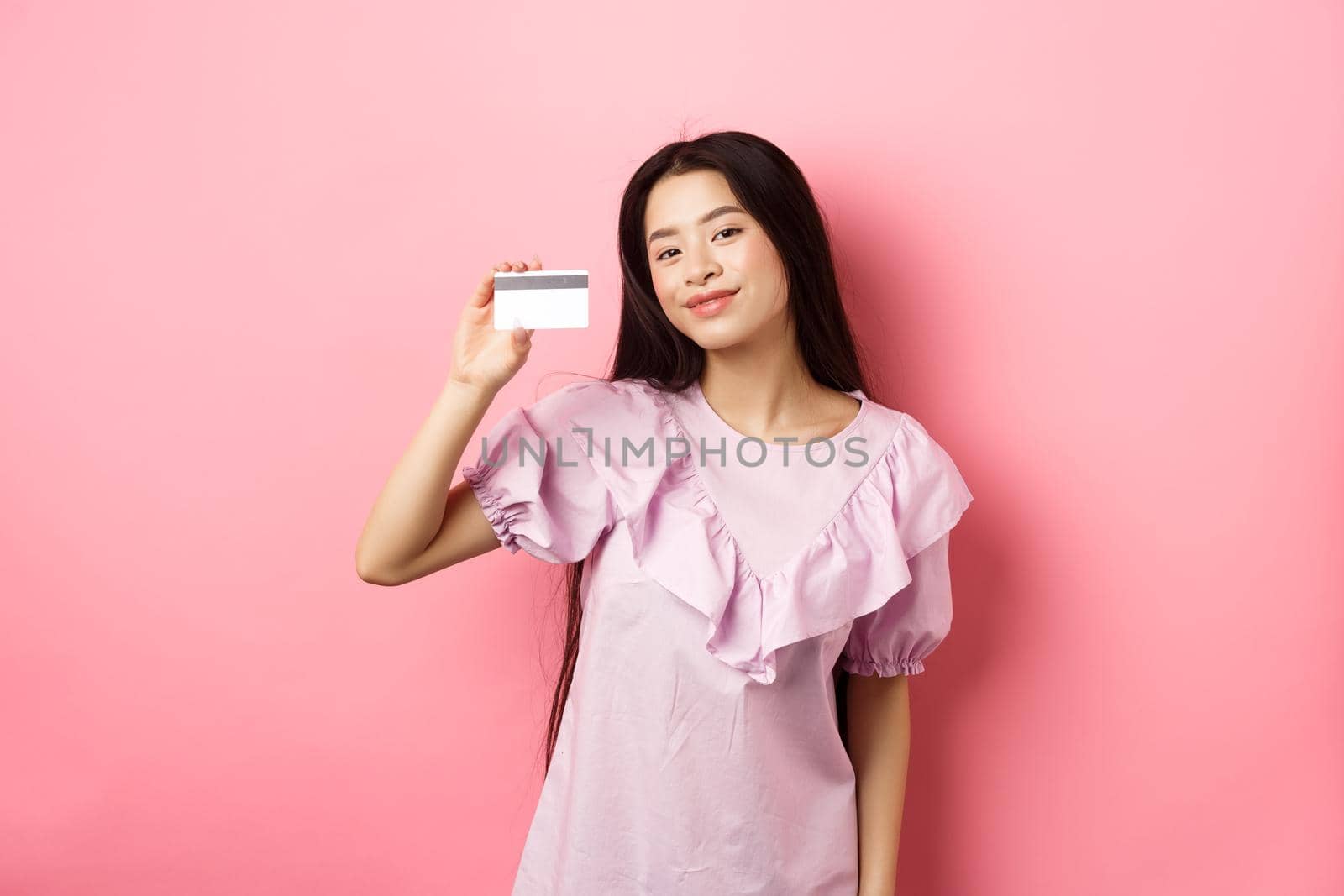 Shopping. Young korean girl in dress showing credit card, smiling satisfied at camera, standing on pink background.