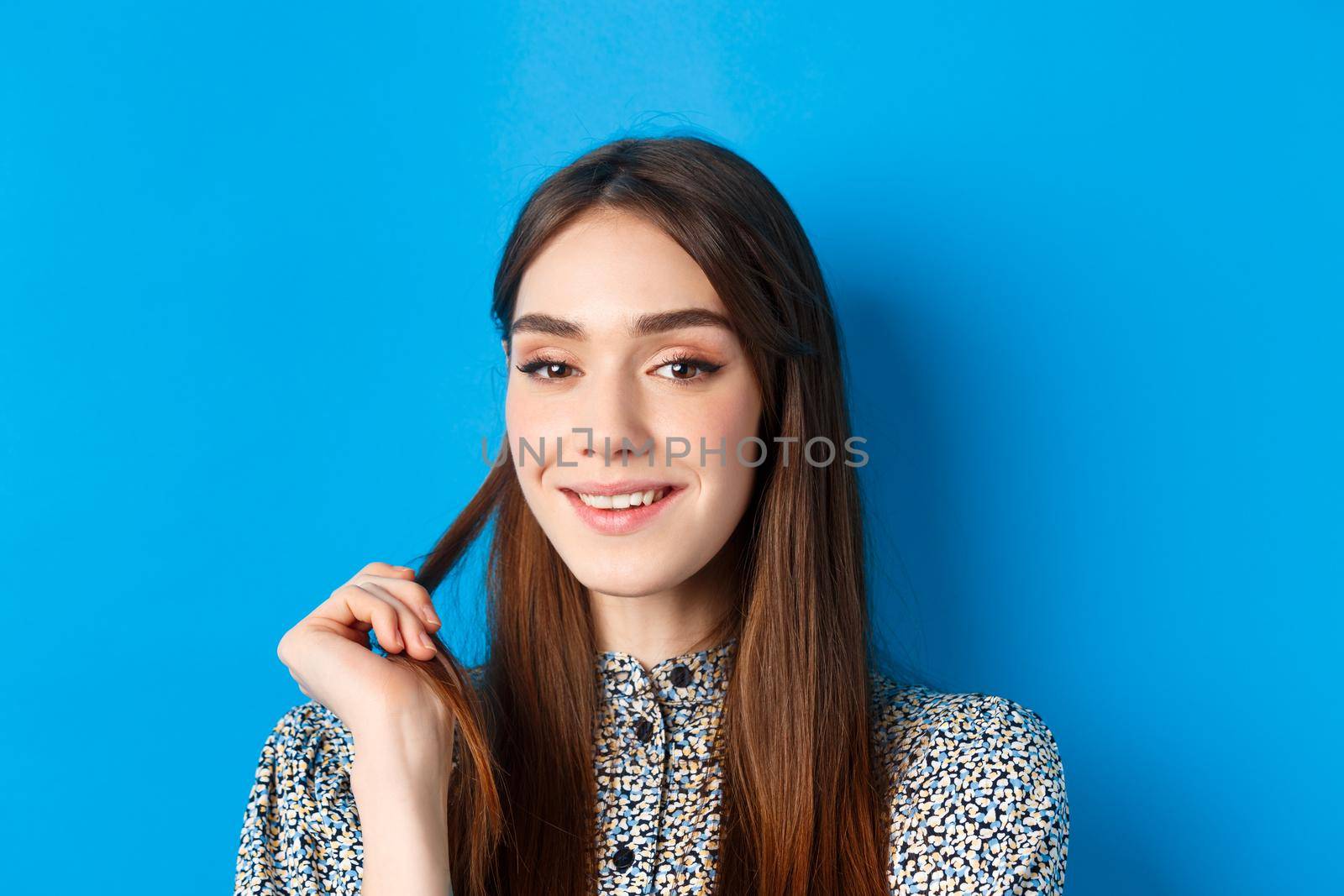 Close-up portrait of flirty young woman with natural beauty, smiling and playing with hair strand, having interesting idea, standing on blue background.